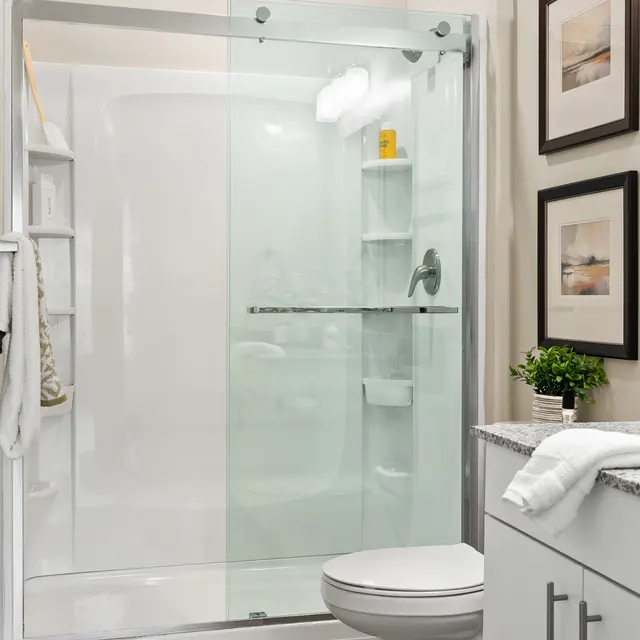 A modern bathroom featuring a glass shower enclosure, toilet, and a small cabinet with decorative frames on the wall.