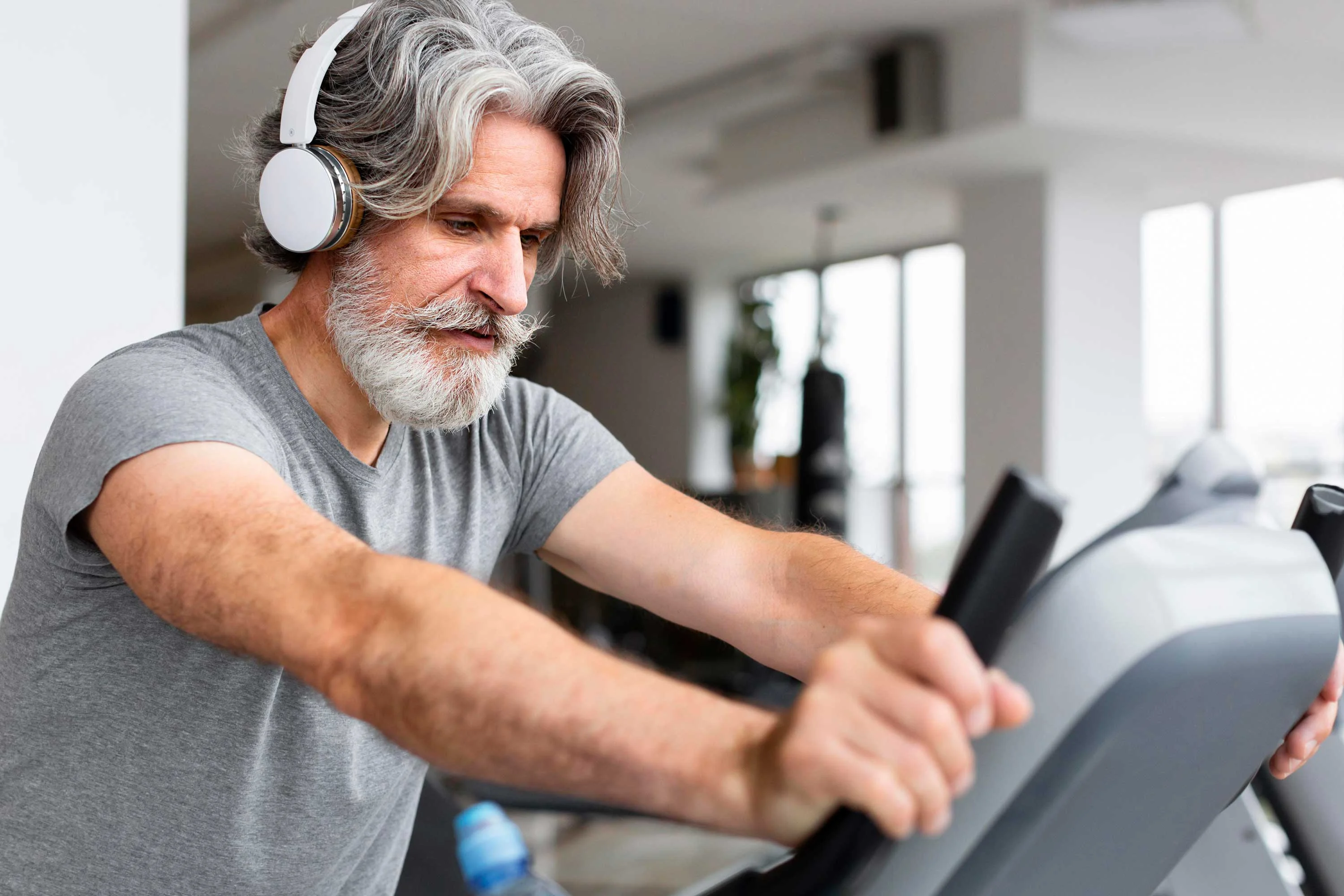 Senior Man Exercising in Gym A senior man with a gray beard and long hair is exercising on an elliptical machine in a modern gym, wearing headphones and focused on his workout.