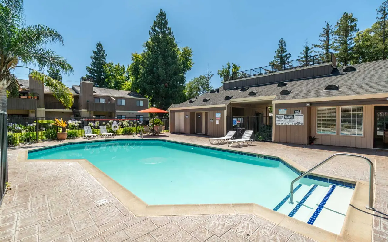A clear blue swimming pool surrounded by lounge chairs and lush green palm trees, with a clubhouse in the background.