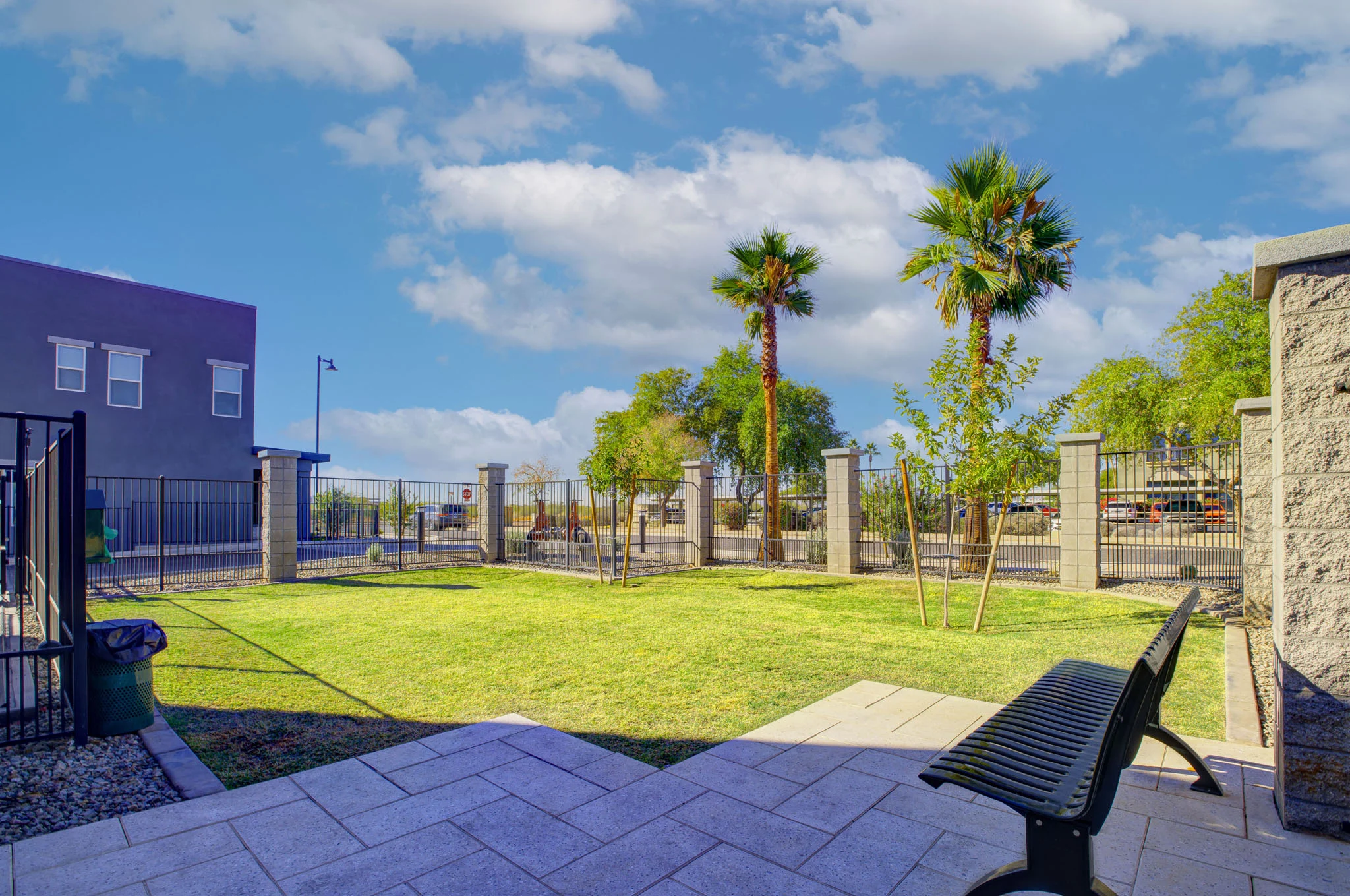 A landscaped area featuring a green lawn with young trees and palm trees, bordered by a fence and a concrete pathway, under a bright blue sky with some clouds.