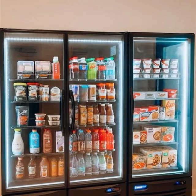 A refrigerator with glass doors showcasing various drinks and snacks inside. The left section has dairy products, the middle section contains a variety of bottled drinks, and the right section holds different types of ready-to-eat meals and snacks.