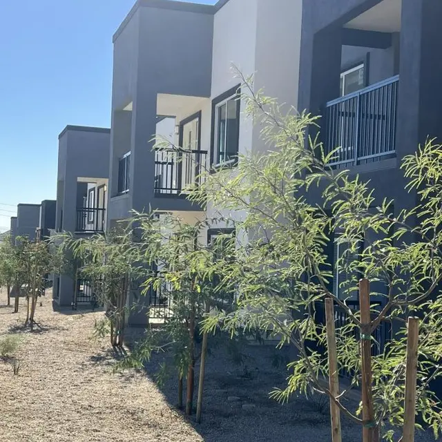 A modern apartment complex with a contemporary architectural design, featuring multiple units with balconies. Young trees are planted in front of the units on a gravel landscape under a clear blue sky.