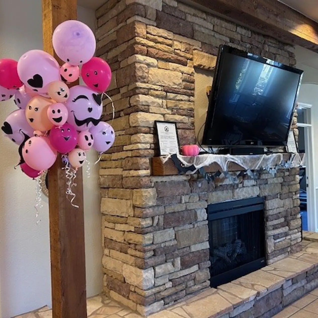 A cluster of pink and lavender balloons with smiley faces attached to the wall next to a stone fireplace, with a TV mounted above.