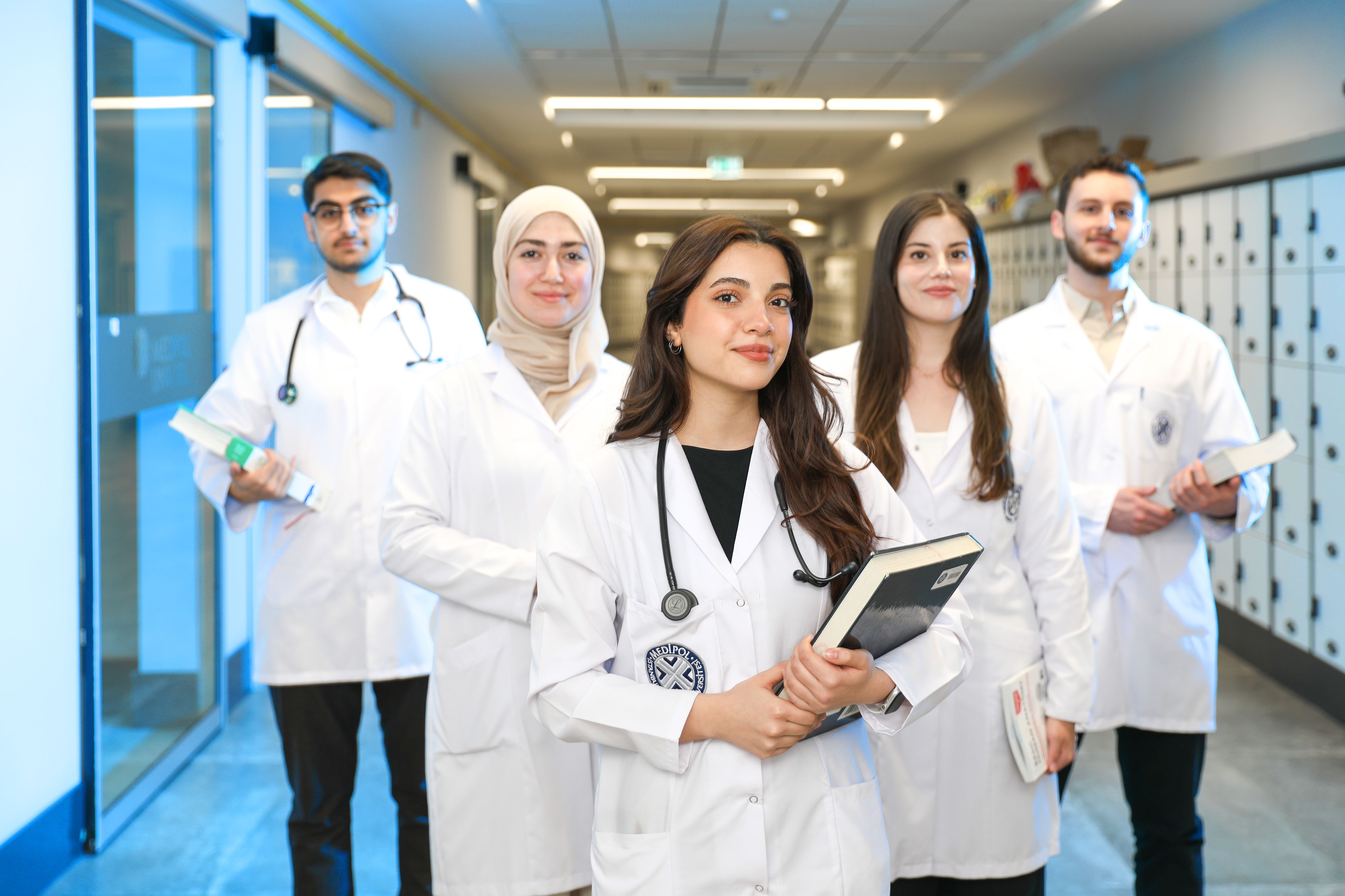 A group of five diverse medical students in white lab coats standing in a hallway, looking confidently at the camera, with lockers in the background.