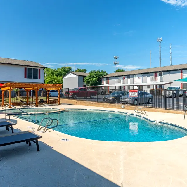 An outdoor pool area at an apartment complex featuring lounge chairs, umbrellas, and a gazebo with a fence in the background and cars parked nearby.