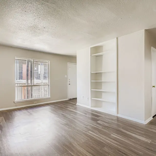 An empty living room in an apartment featuring light-colored walls, wooden floors, a large window, and a built-in shelf.
