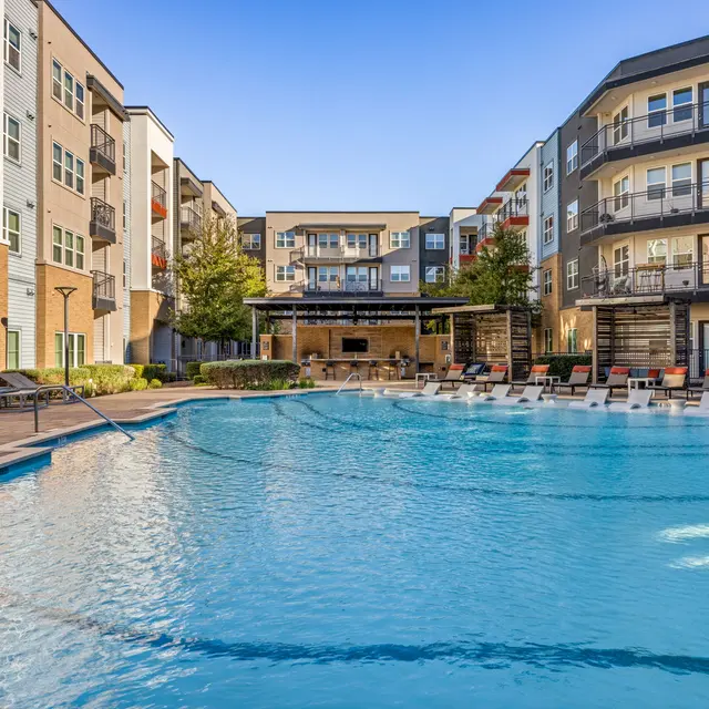 A clear swimming pool surrounded by modern apartment buildings with loungers and a covered seating area.