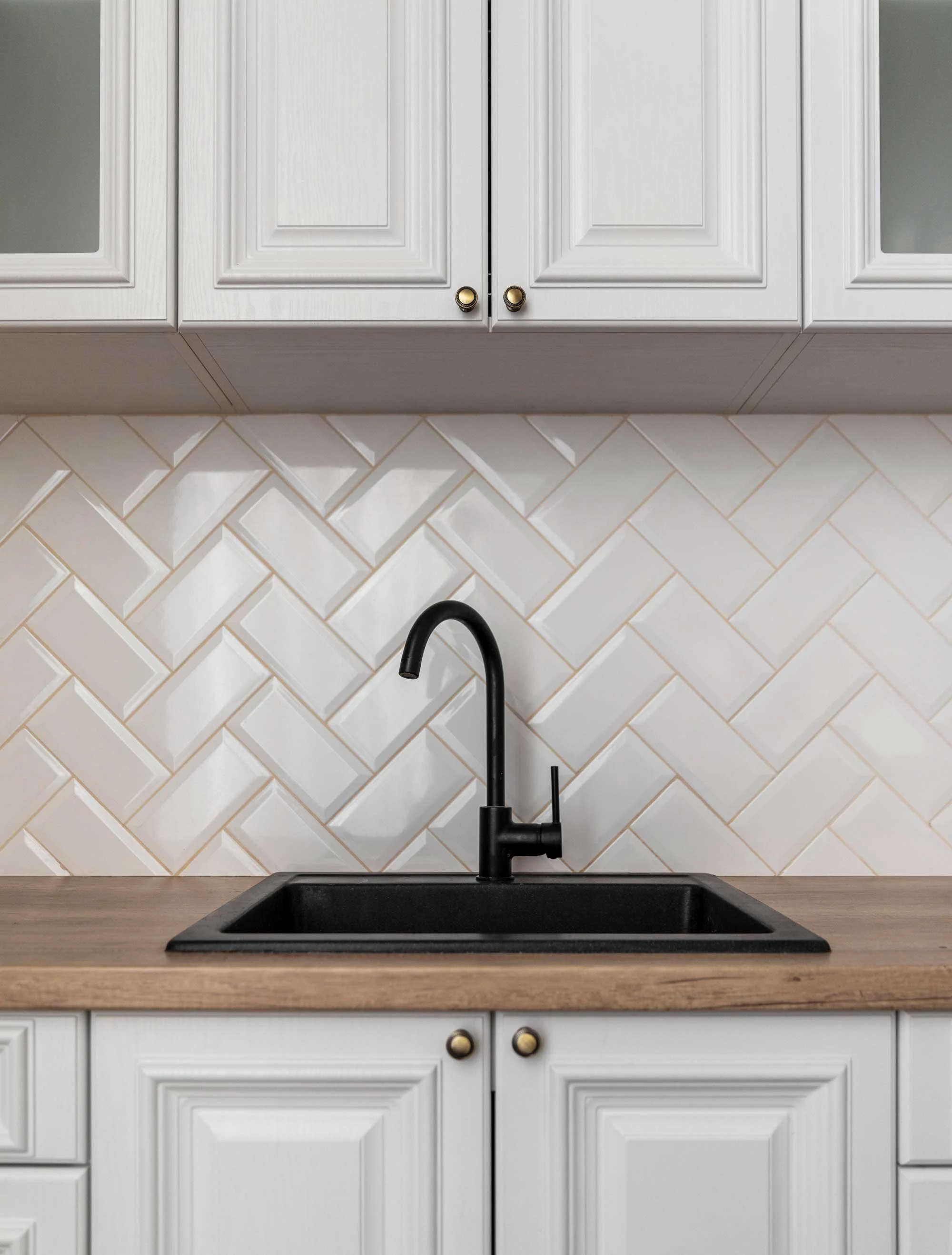 A modern kitchen sink with a black faucet and a black basin installed on a wooden countertop. The background features white cabinets with brass knobs and a herringbone tile backsplash.
