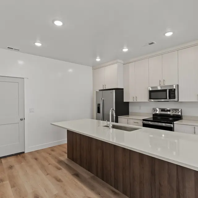 A modern kitchen featuring light-colored cabinetry, stainless steel appliances, and a large central island with a sink. The floor is made of wooden planks, and the walls are painted white, creating a bright and airy feel. There is a closed door visible on the left side.
