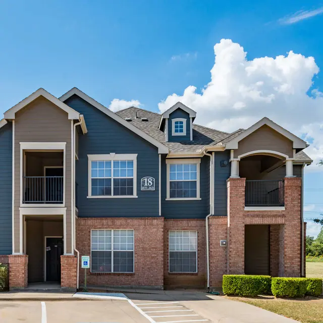 A two-story apartment building with a combination of blue siding and red brick, featuring balconies and trees in the foreground under a clear blue sky.