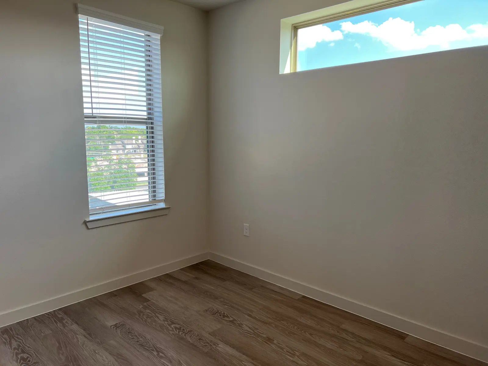 A vacant room featuring light-colored walls, a window with blinds, and a wooden floor. A small window is visible at the top, allowing natural light to illuminate the space.