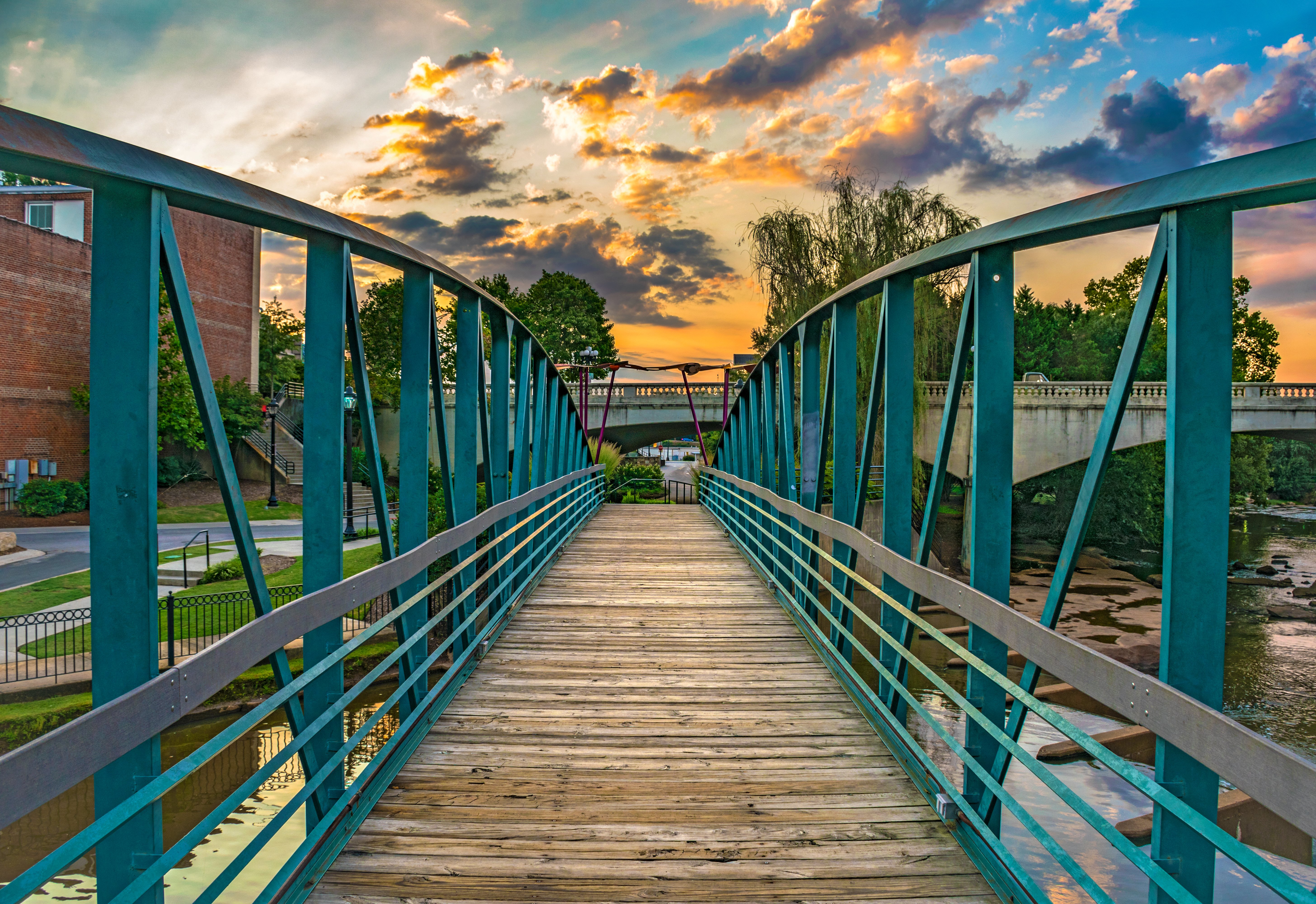 Scenic Footbridge at Sunset A scenic view of a wooden footbridge with teal supports, leading towards a sunset with colorful clouds. The bridge is surrounded by greenery and water, creating a tranquil atmosphere.