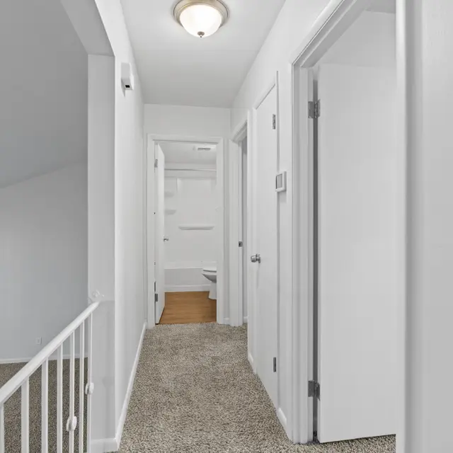 Interior view of a hallway with white walls and carpet, leading to doors and a small bathroom at the end.