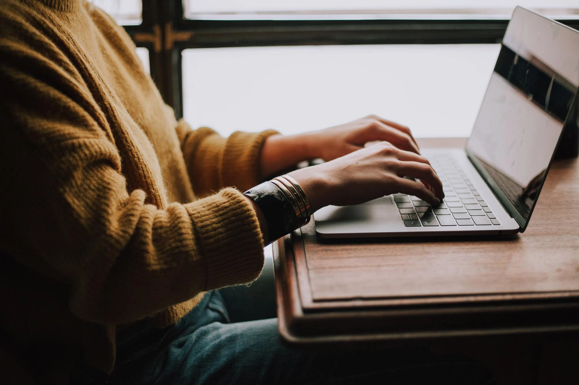 A person typing on a laptop at a wooden table, wearing a yellow sweater and jeans, with sunlight streaming in through a window.