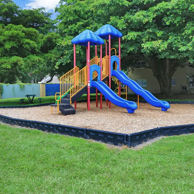 Colorful playground structure with two slides, surrounded by grass and trees.