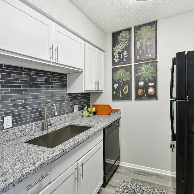 A modern kitchen featuring sleek white cabinetry, a granite countertop, and a stylish backsplash. The kitchen includes a black refrigerator and stove, with decorative fruit-themed artwork on the wall.