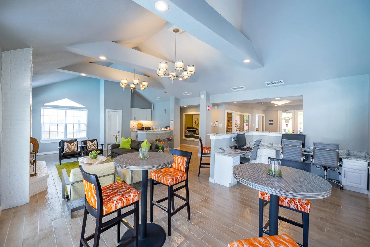 A spacious modern living area featuring various seating arrangements and bright decor. The room has a high ceiling with stylish lighting and large windows providing natural light. The furniture includes circular tables with colorful chairs and a cozy seating area.