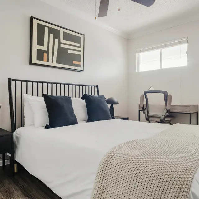 A modern bedroom featuring a queen-sized bed with white linens and navy blue cushions. There's a wooden nightstand with a black lamp, a work desk with a chair in the background, and abstract wall art above the bed. The room has a large window letting in natural light.