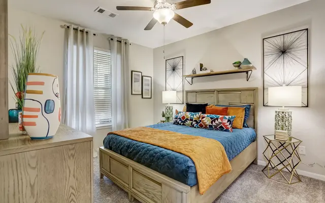 A cozy bedroom featuring a large bed with colorful pillows, an orange throw blanket, two bedside lamps, and decorative wall art. The room has light curtains and a fan overhead.