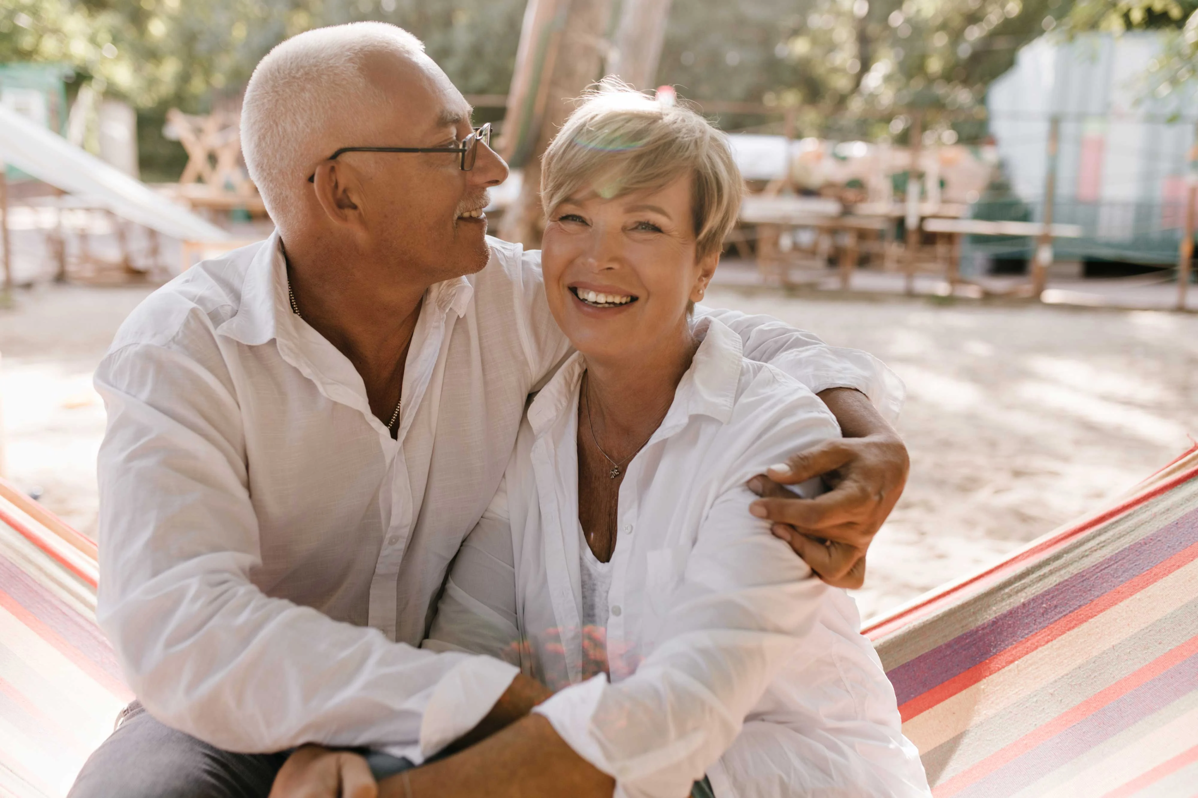 A happy couple sitting together in a hammock, smiling at each other. The man has short white hair and glasses, wearing a white shirt. The woman has short, blonde hair and is also dressed in a white shirt. The background shows a tranquil outdoor setting with trees and soft sunlight.