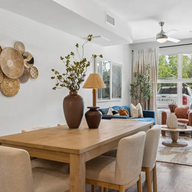 Stylish Dining and Living Room A cozy dining area with a wooden table and beige chairs, decorated with a vase. The living room features a blue couch and a stylish rug, with decorative wall hangings.