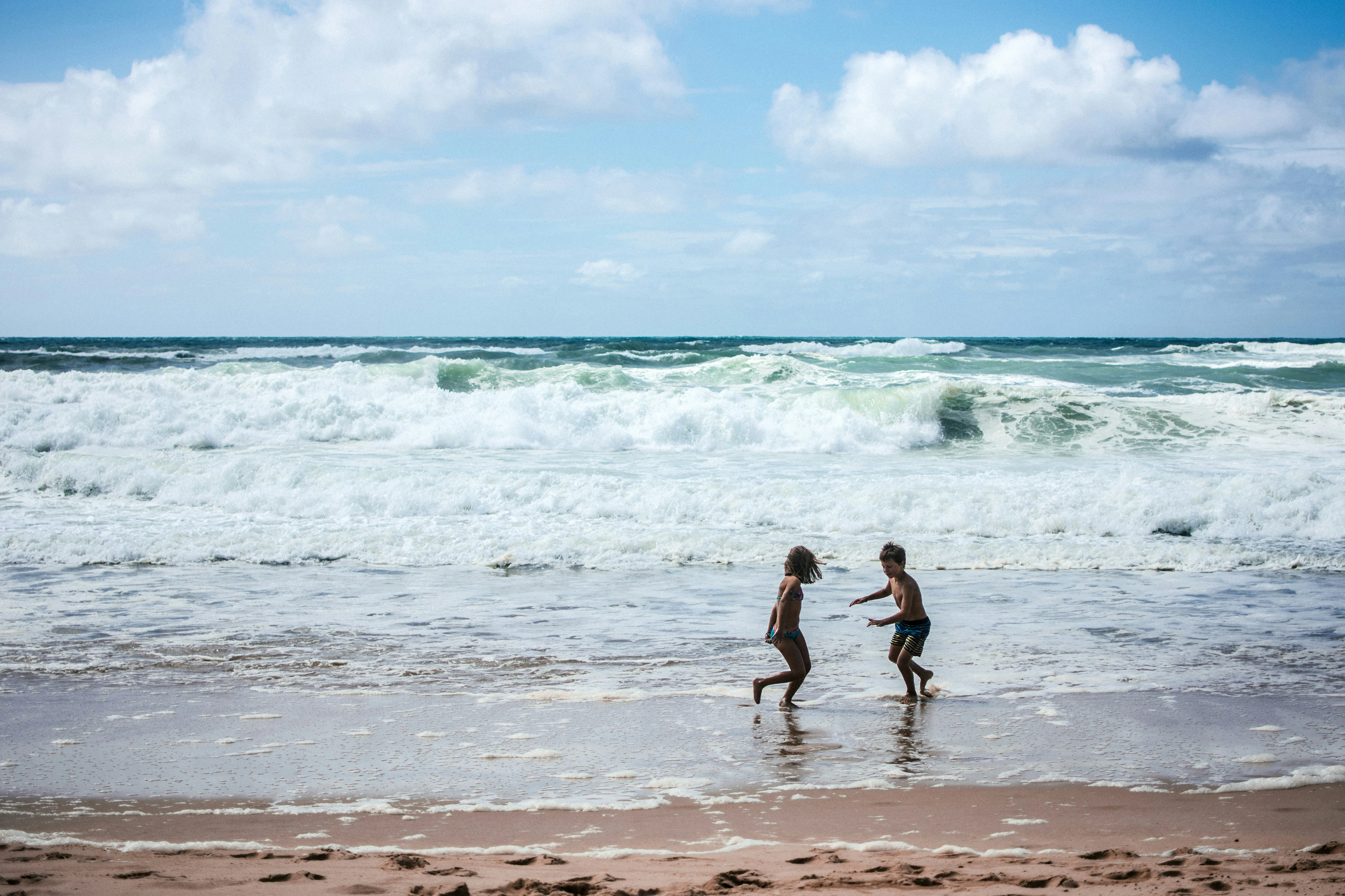 Two children playing in shallow water on a beach, with waves crashing in the background and cloudy skies overhead.