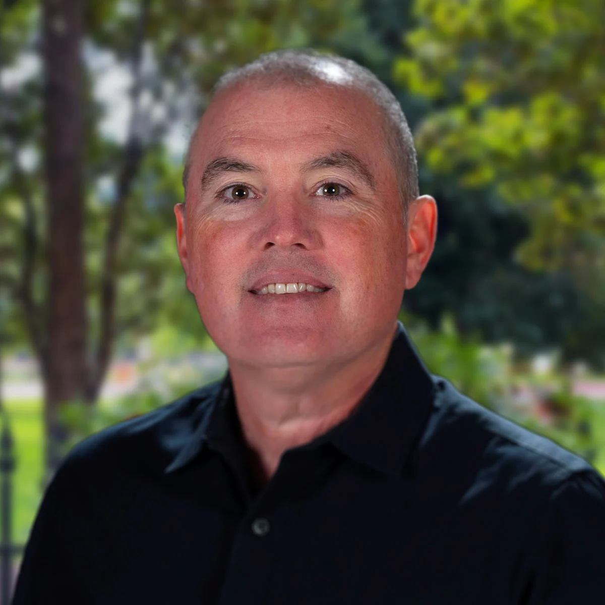 A man with short gray hair and a black shirt, smiling against a blurred green background with trees.