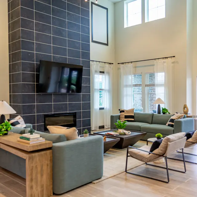 A spacious modern living room with light-colored walls, a large fireplace with a dark tiled accent wall, a flat-screen TV, a comfortable sectional sofa, and stylish chairs. The decor includes greenery and contemporary light fixtures, creating a bright and inviting atmosphere.