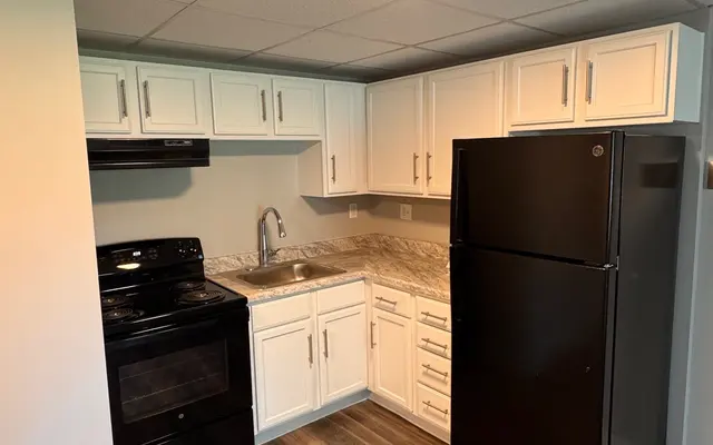 Modern Kitchen Design A small modern kitchen featuring black appliances including an oven and refrigerator, white cabinets, and a granite countertop. The floor has a wooden texture and the ceiling has a grid design with ambient lighting.