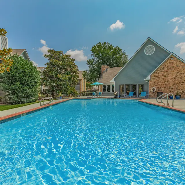 A spacious swimming pool surrounded by greenery and a residential building, with clear blue water reflecting the sky.