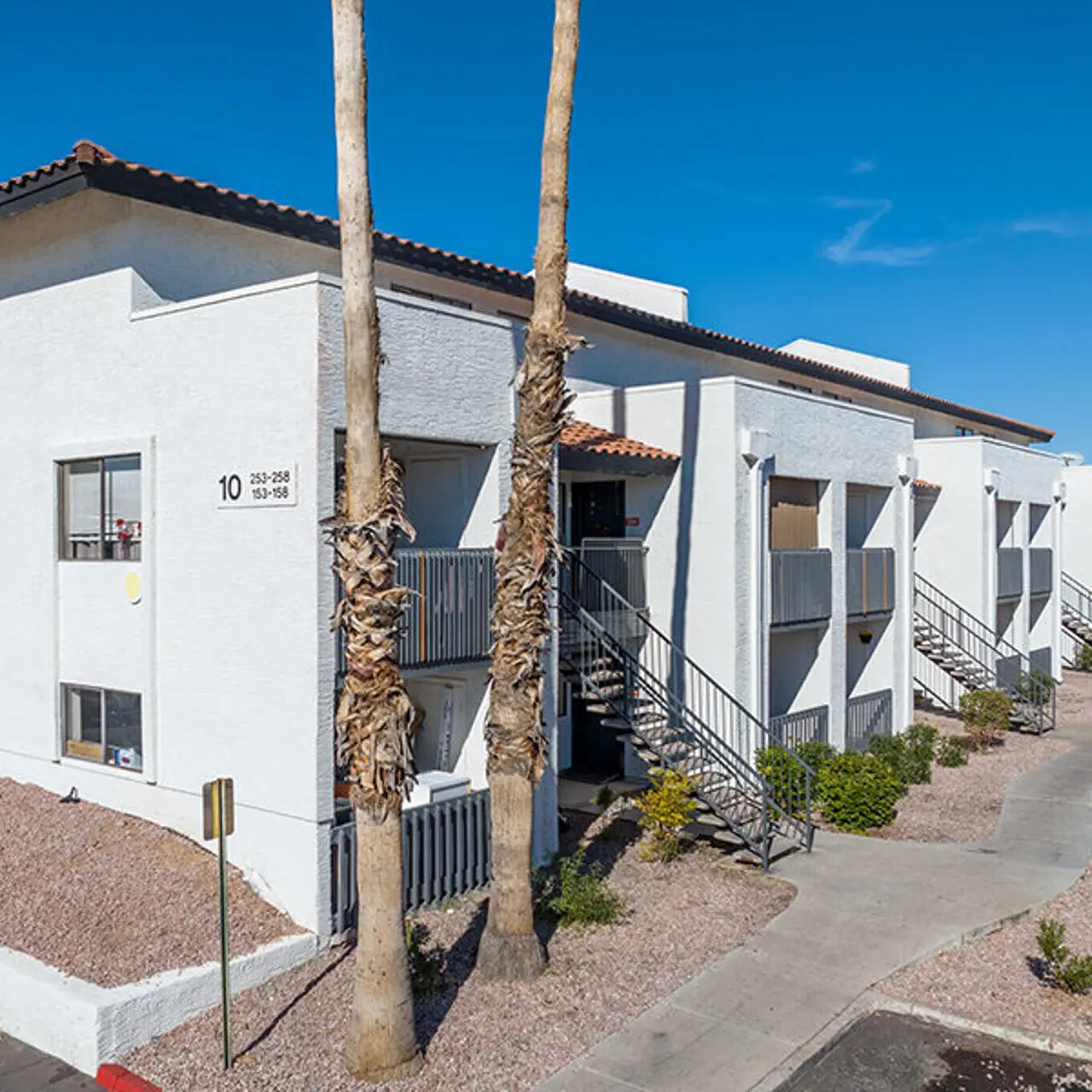 Exterior view of a two-story apartment complex featuring white walls, balconies, and palm trees.