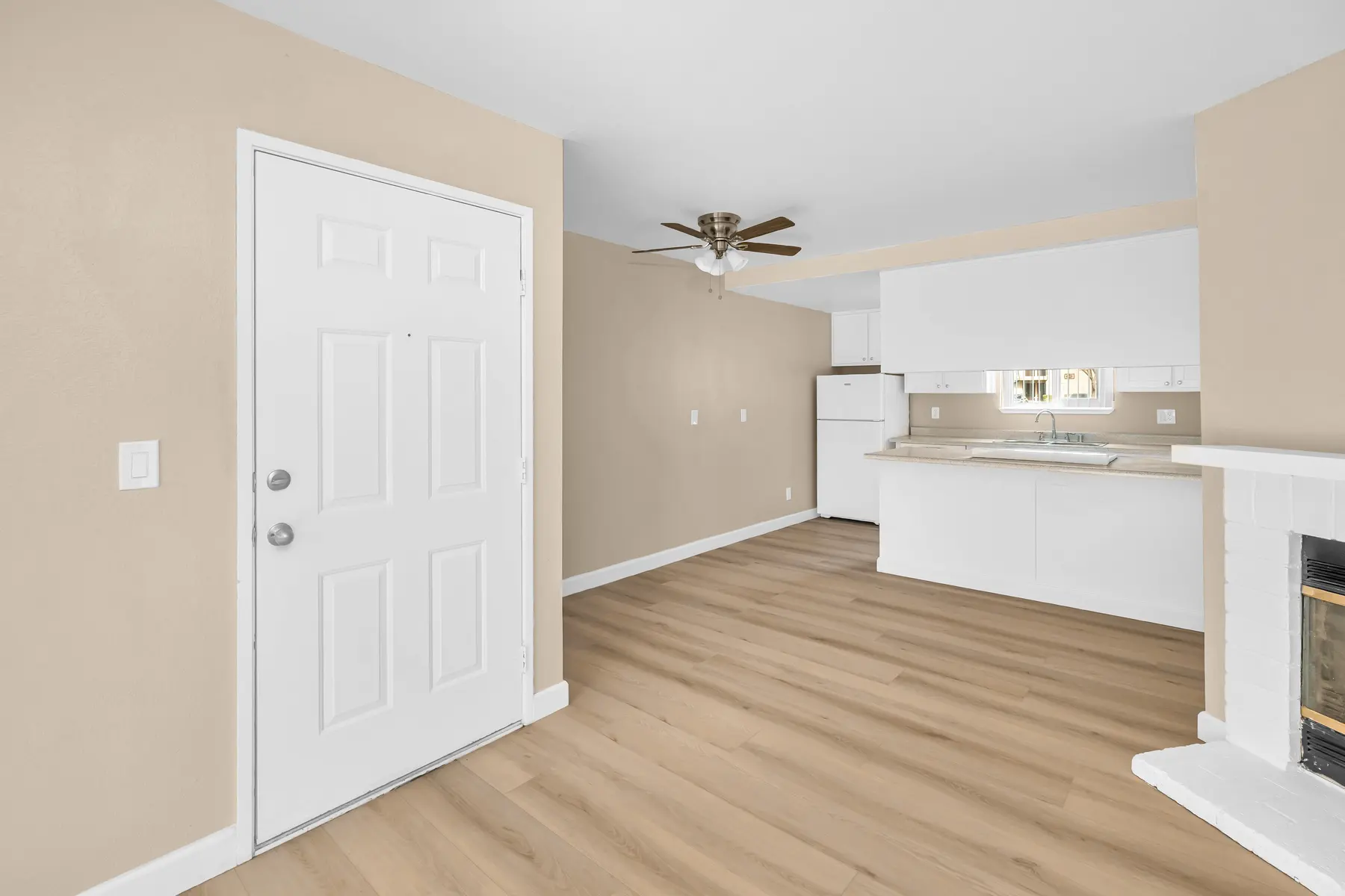 Interior view of a modern apartment showing an open space with light wood flooring, beige walls, a white door, and a simple kitchen area visible in the background.