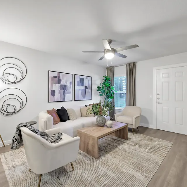 A modern living room featuring a light-colored sofa, two armchairs, and a wooden coffee table. The room has abstract wall art, a decorative floor lamp, and a green plant. A door leads outside, and there are light curtains by the window.