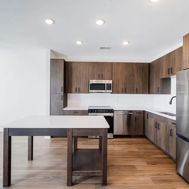 A modern kitchen featuring wooden cabinetry, stainless steel appliances, and a white countertop island. The space is well-lit with recessed lights and has a contemporary design.