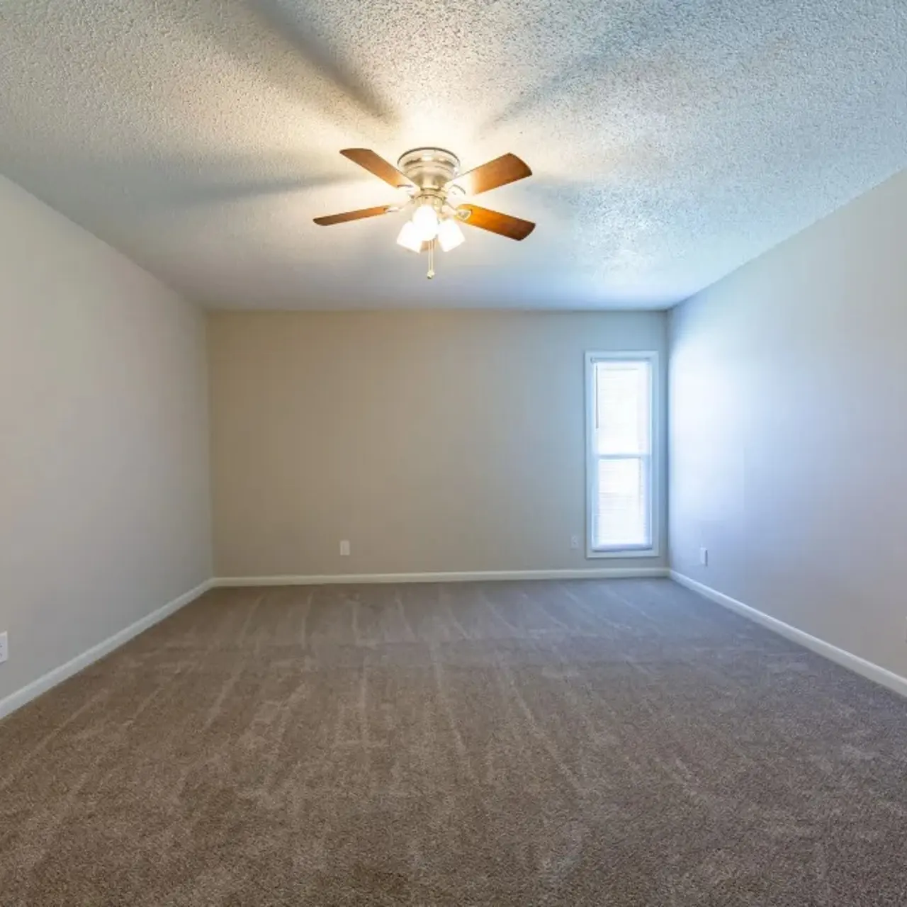 An empty room featuring beige walls and a ceiling fan. A window on the right allows natural light to enter.