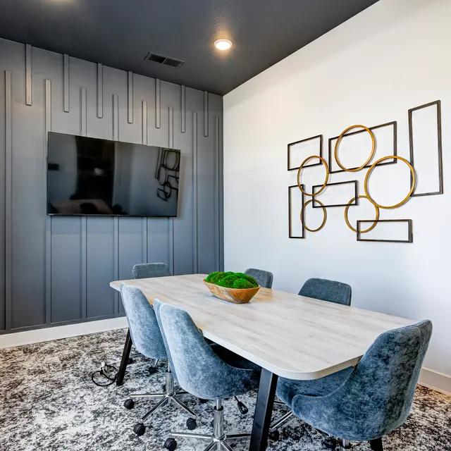 A modern meeting room featuring a round table with a potted plant in the center, surrounded by plush blue chairs. A wall-mounted television is visible on the dark accent wall, while geometric wall art adds decor to the white wall opposite.