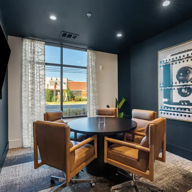 A modern meeting room with a round table surrounded by four leather chairs. There is a large window with light curtains allowing natural light to enter, and a piece of framed wall art featuring a film reel design.