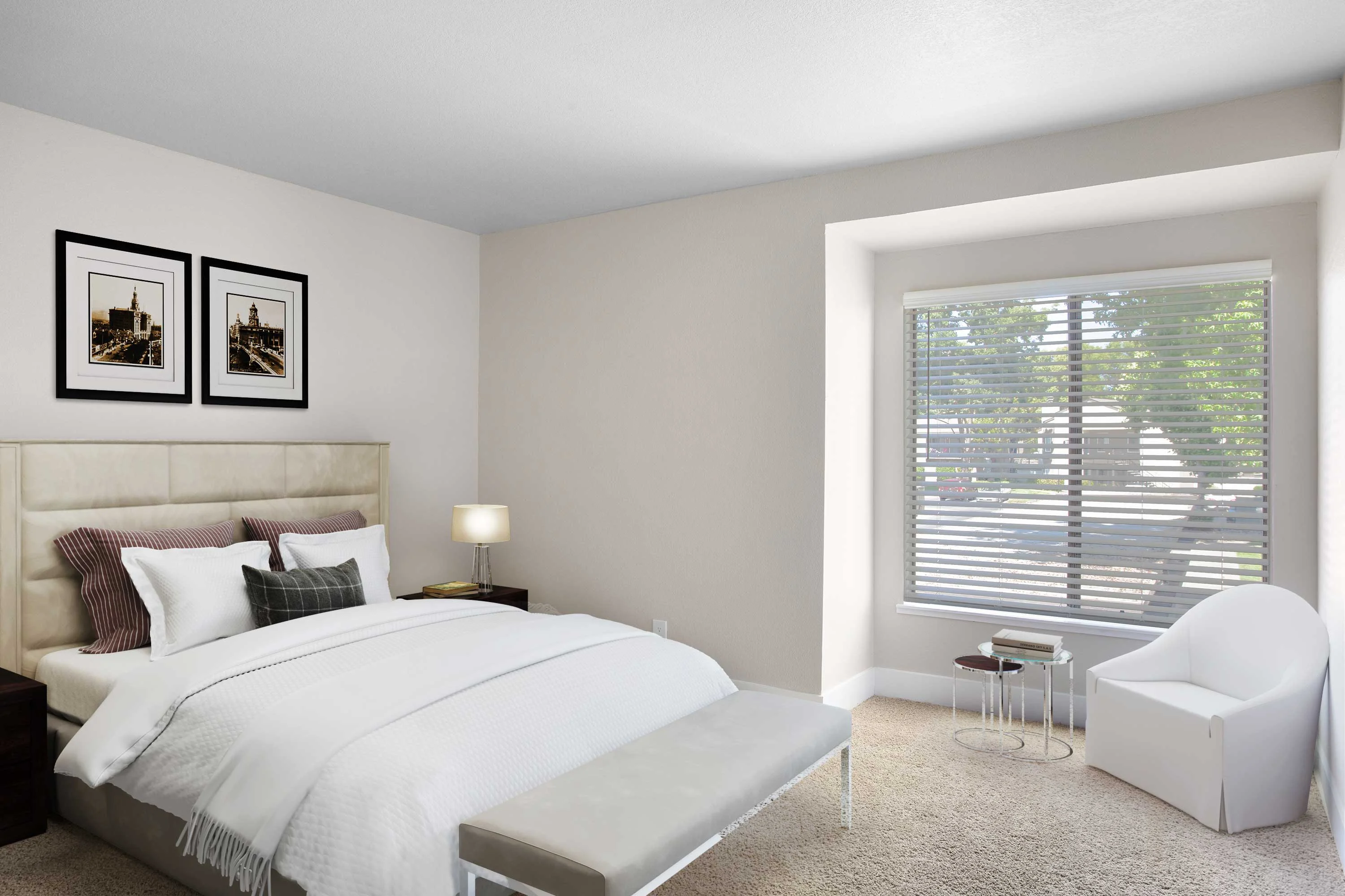 Modern Bedroom Interior A modern bedroom featuring a beige headboard bed with white bedding, accompanied by a bench at the foot and a small side table with a lamp. There are two framed pictures on the wall, and a large window with blinds allows natural light to fill the room.
