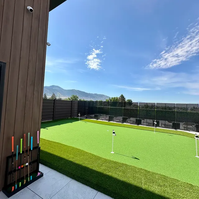 A well-maintained outdoor putting green with flag poles, surrounded by a fence, under a bright blue sky with a few clouds.