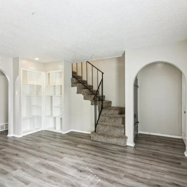 A spacious living room with wooden flooring and a staircase leading to an upper level. There are built-in shelves on the left side and archways on both sides of the room.