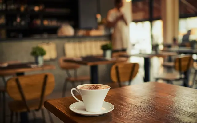 A cup of coffee on a wooden table in a cozy cafe setting, with blurred background showing indoor plants and a barista. The decor exudes a warm, inviting atmosphere.