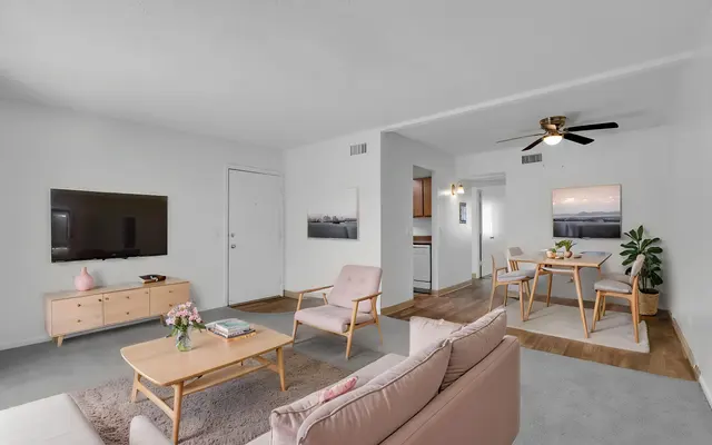 A modern living room with light-colored furniture, a television mounted on the wall, and a dining area visible in the background. The space is bright and airy with a neutral color palette.
