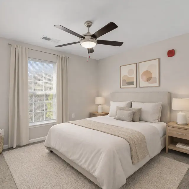 A modern bedroom featuring a queen bed with light gray bedding, side tables with lamps, and decorative wall art. A window with white curtains allows natural light, and a woven basket is positioned next to the bed.