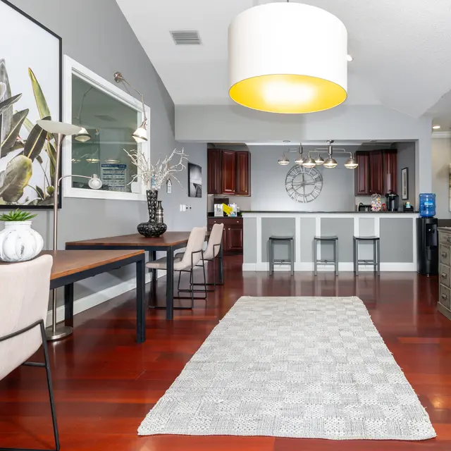 A modern living area featuring a dining and workspace setup. There are two chairs at a light wooden table and a long rug stretching across the floor. In the background, a kitchen with dark wooden cabinets and a clock on the wall is visible. A large circular light fixture hangs above the room, enhancing the modern aesthetic.
