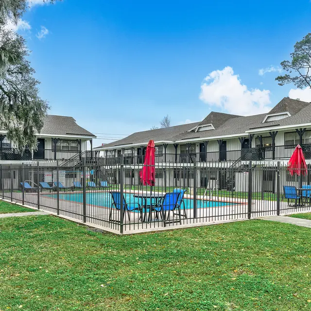 A view of a courtyard in an apartment complex featuring a fenced pool area with lounge chairs and umbrellas, surrounded by two-story buildings.