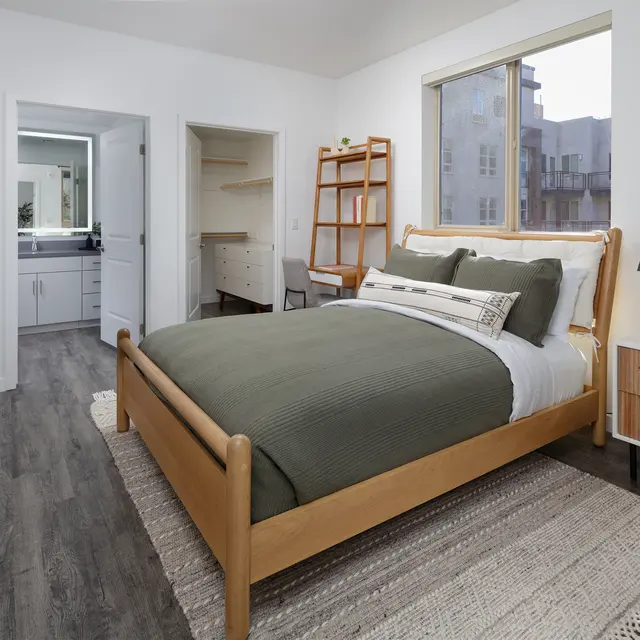 A modern bedroom featuring a wooden bed with a green comforter, accompanied by a nightstand and a wool rug. A mirror is visible on the wall, and a bathroom is accessible through an open door.