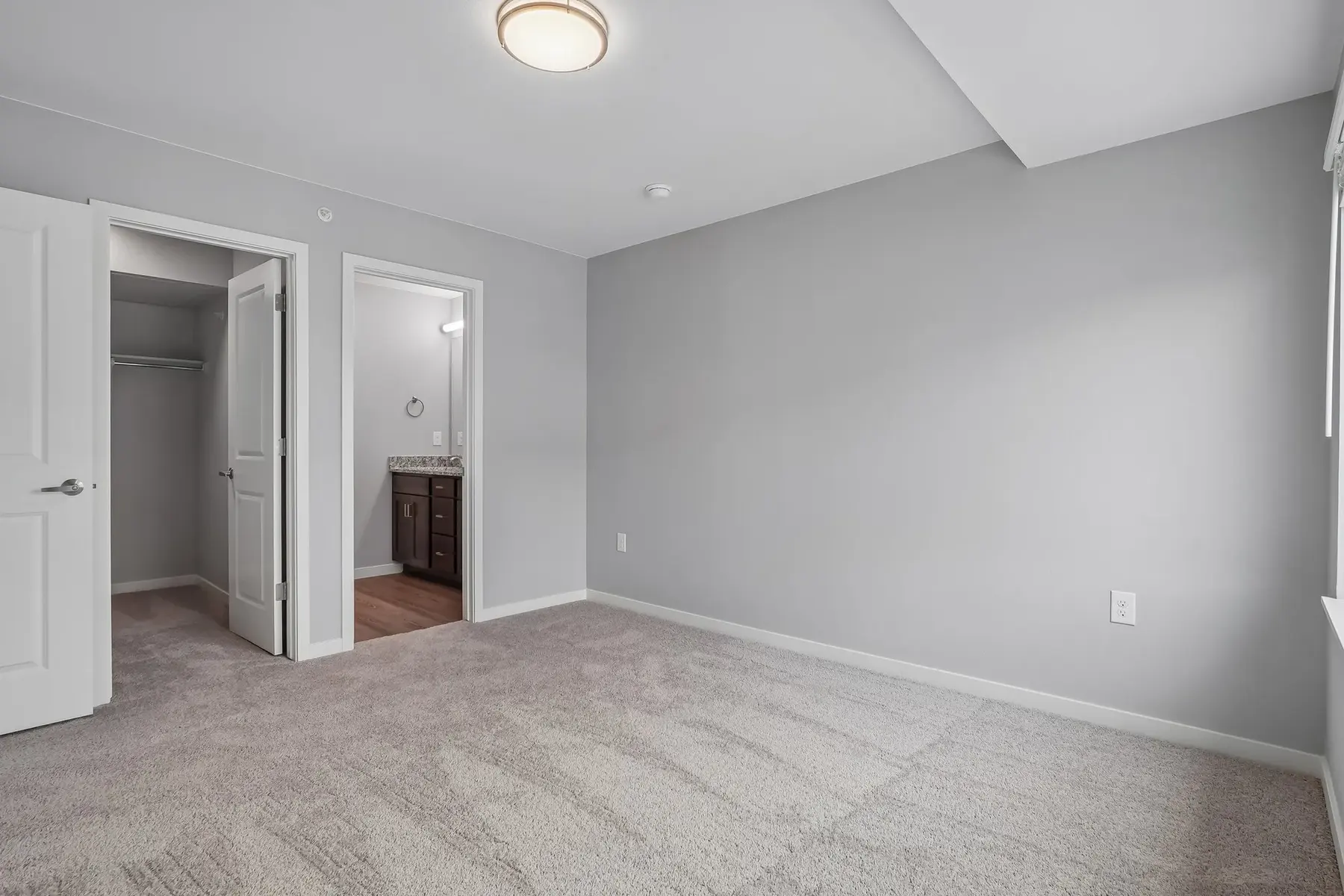 A modern bedroom with gray walls and carpeting, featuring an open closet and a doorway leading to a bathroom or living area.