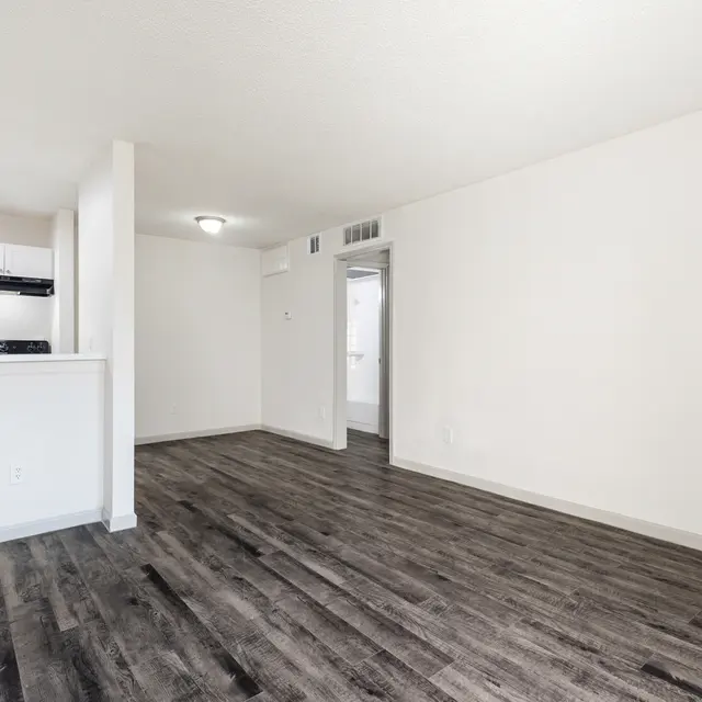 An empty, bright room with light hardwood flooring and white walls. The kitchen is partially visible with a black stove and white cabinetry.