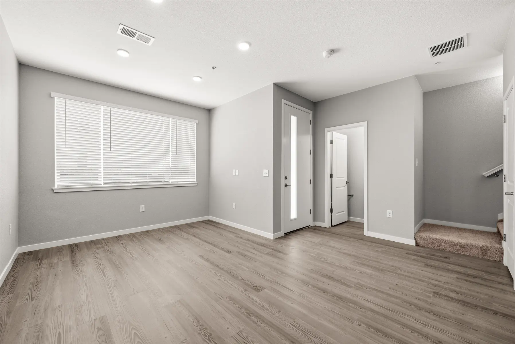 Empty Modern Living Room A spacious empty living room with light gray walls and wooden flooring, featuring large windows with blinds, a door leading to another room, and stairs in the background.