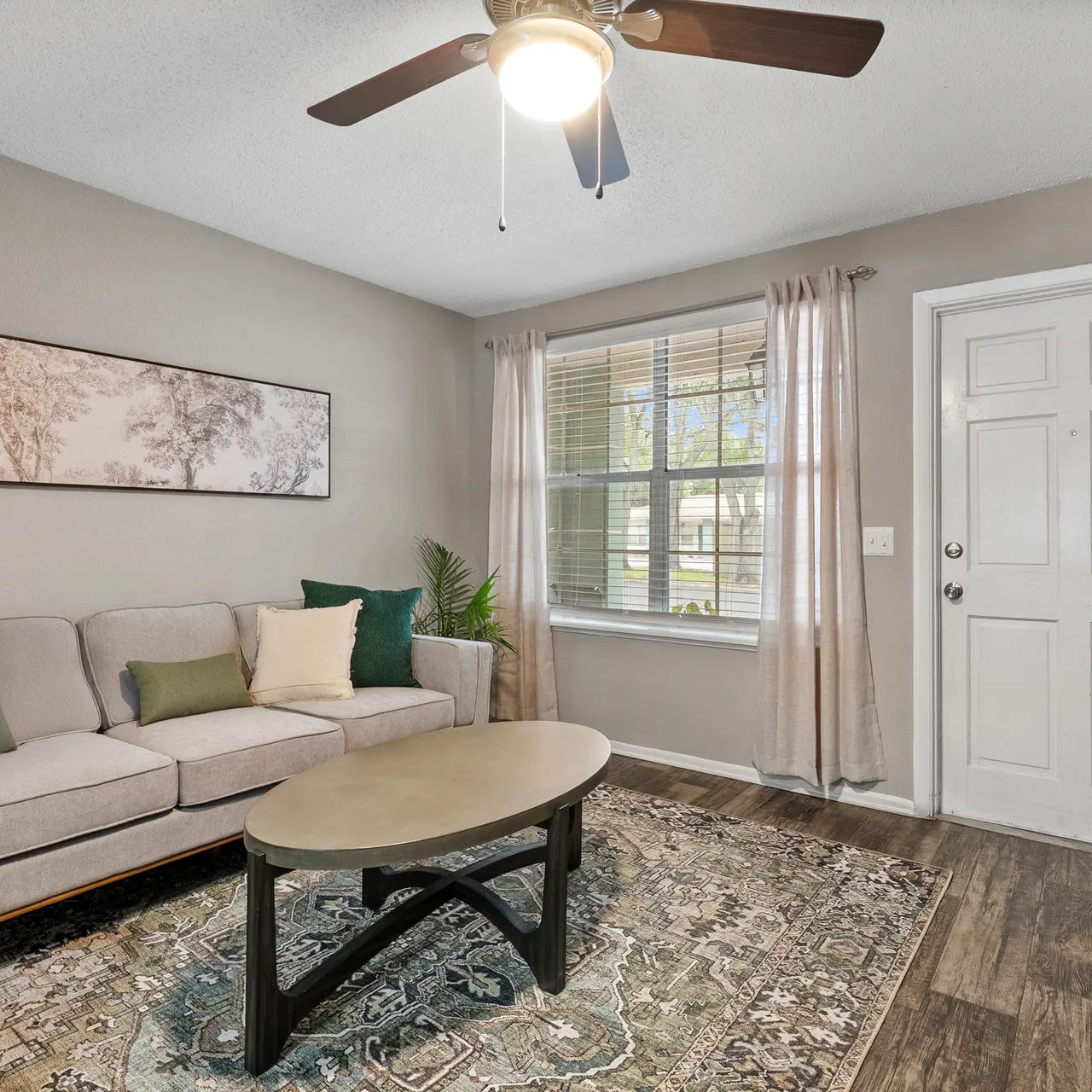 Cozy living room with a light gray sofa, decorative pillows, a round coffee table, and a patterned rug. Natural light comes through the window, and there's a door leading outside.